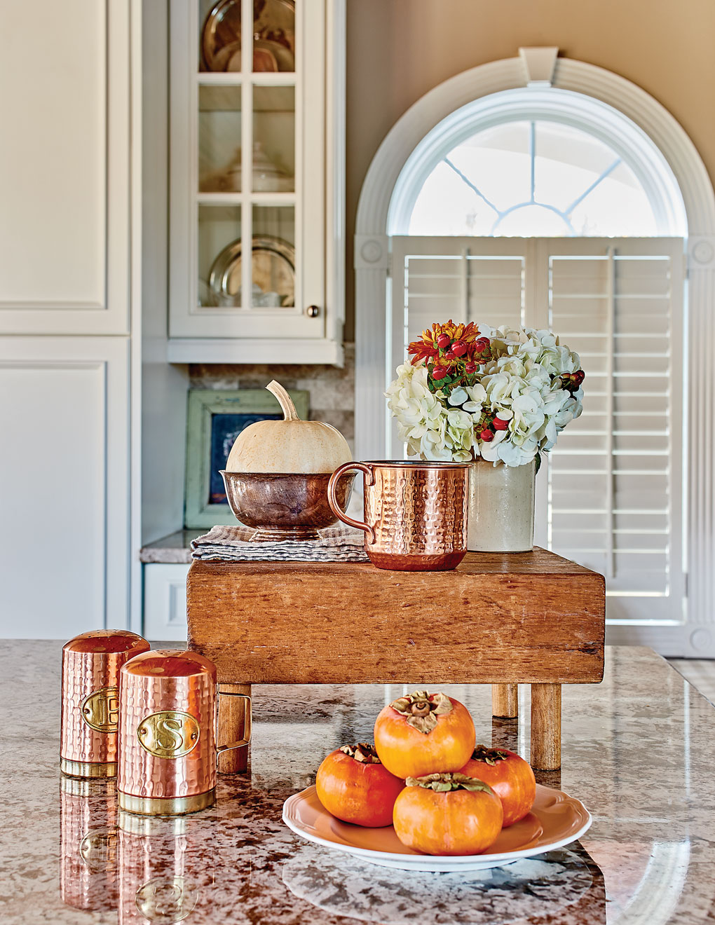 Kitchen view of a granite countertop covered with wooden butcher block display and copper accents alongside a plate of bright orange persimmons. 