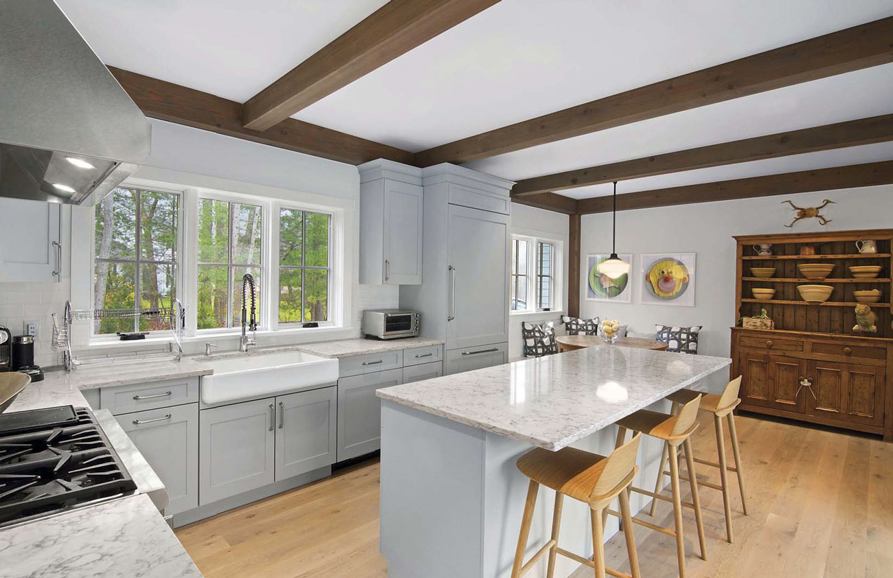 Large gray toned open-concept kitchen with bar/island seating and over looking a dining nook with wooden exposed beams and a large wooden hutch.