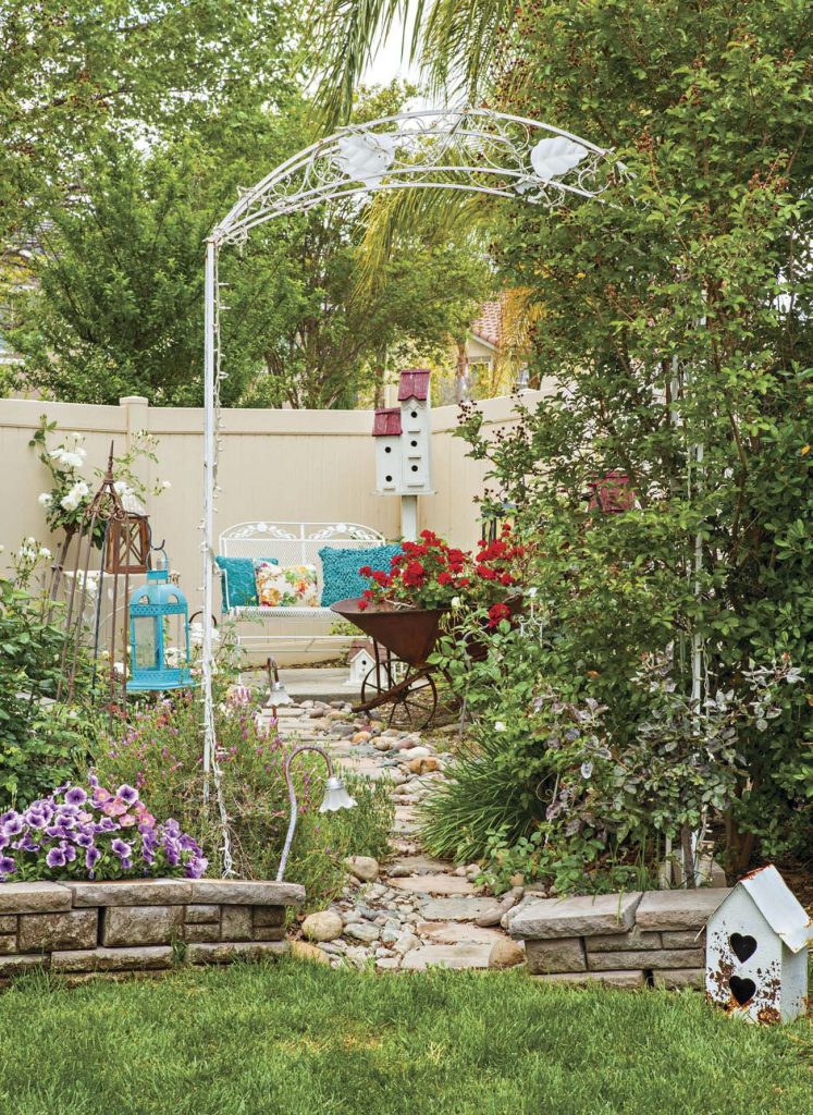 White metal arbor leading to a stone path to an outdoor sitting area with a rustic wheelbarrow filled with red geraniums.