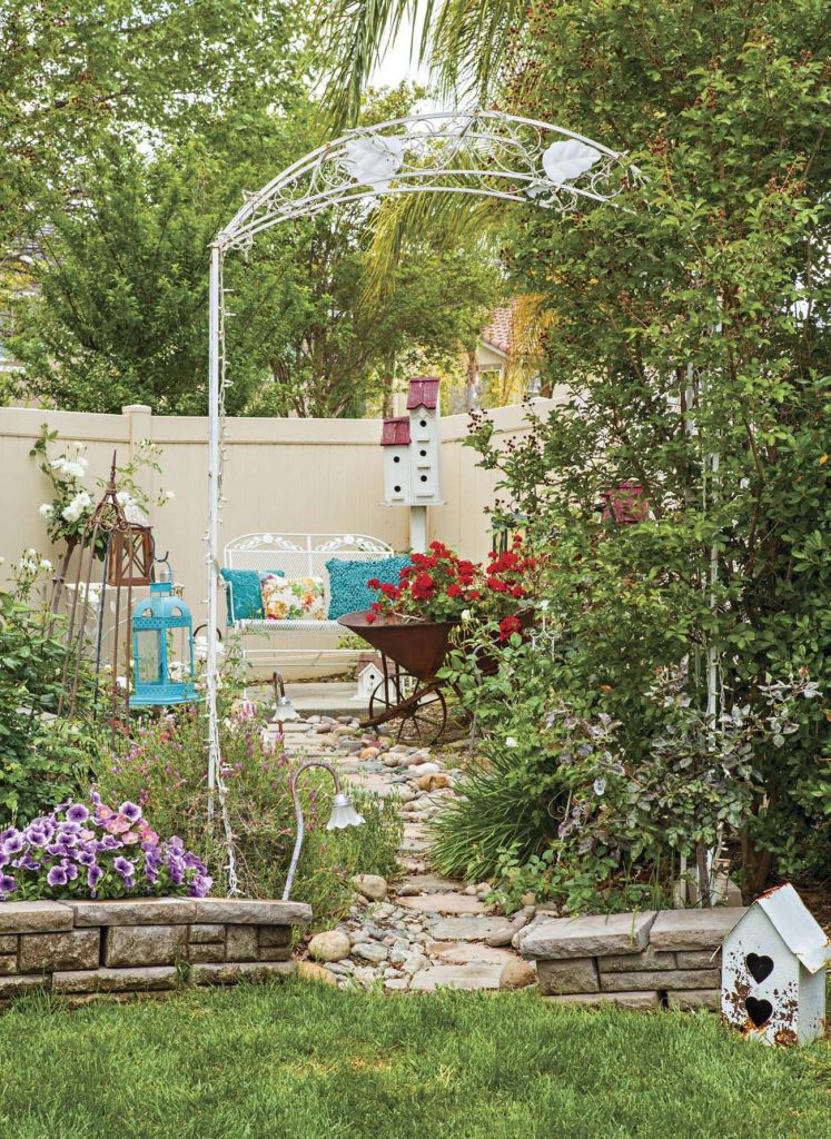 White metal arbor leading to a stone path to an outdoor sitting area with a rustic wheelbarrow filled with red geraniums. 