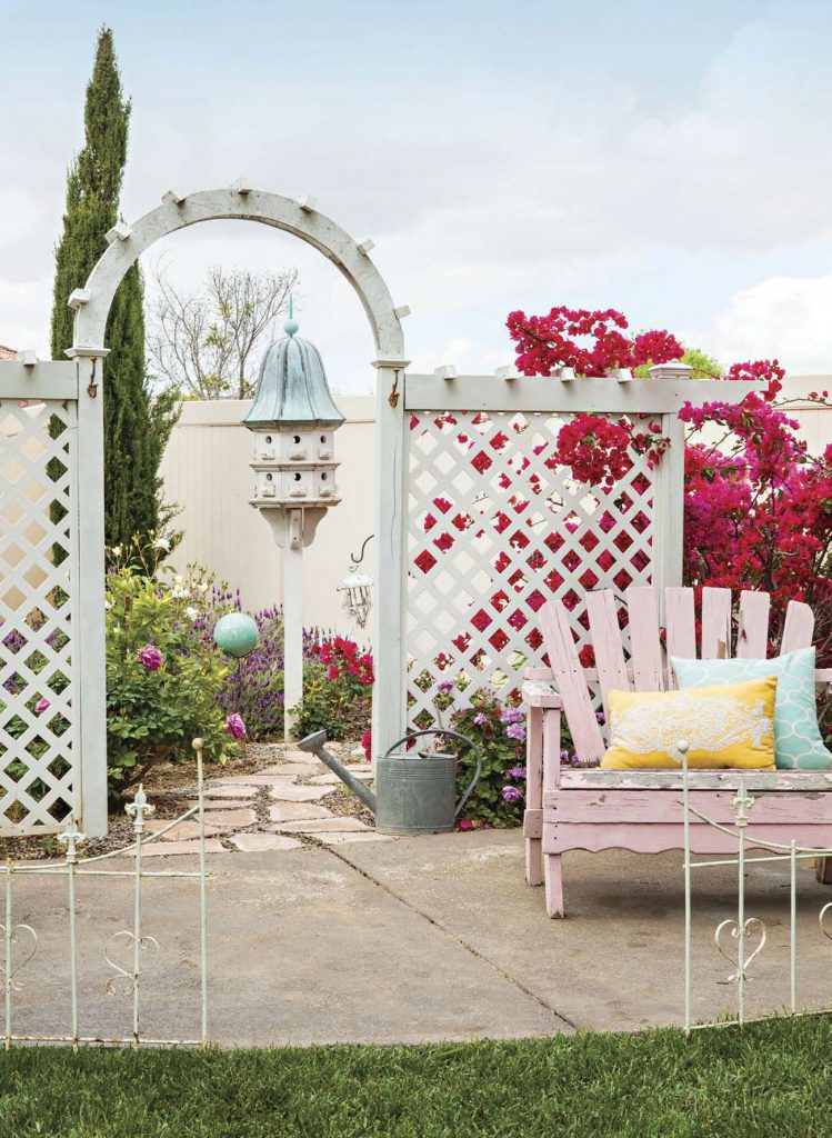 White wooden latticed arbor with climbing pink bougenvilla and a rustic pink adirondak chair in a backyard.