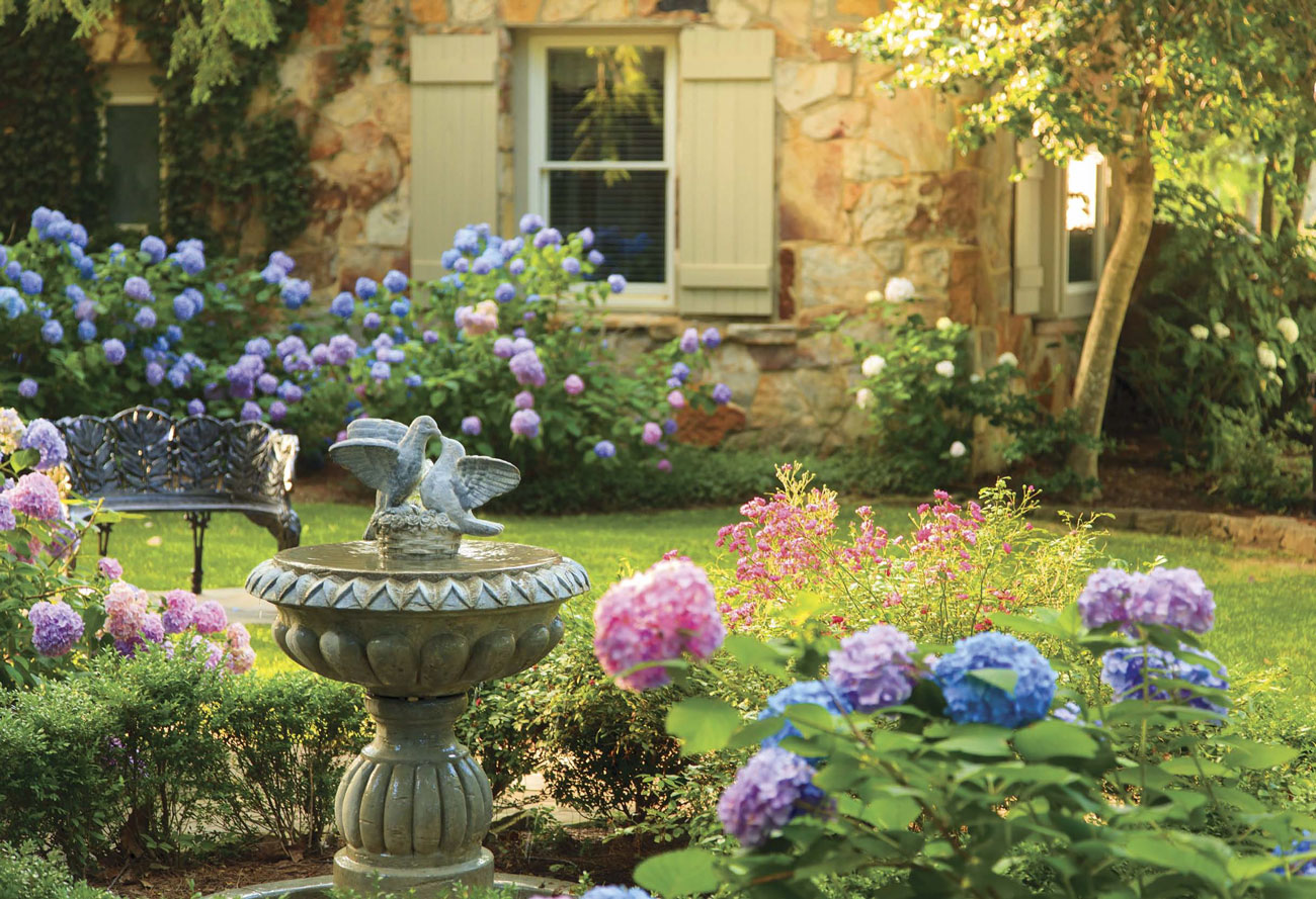 Concrete bird bath surrounded by pink, purple and blue hydrangeas and a wrought iron garden bench. 