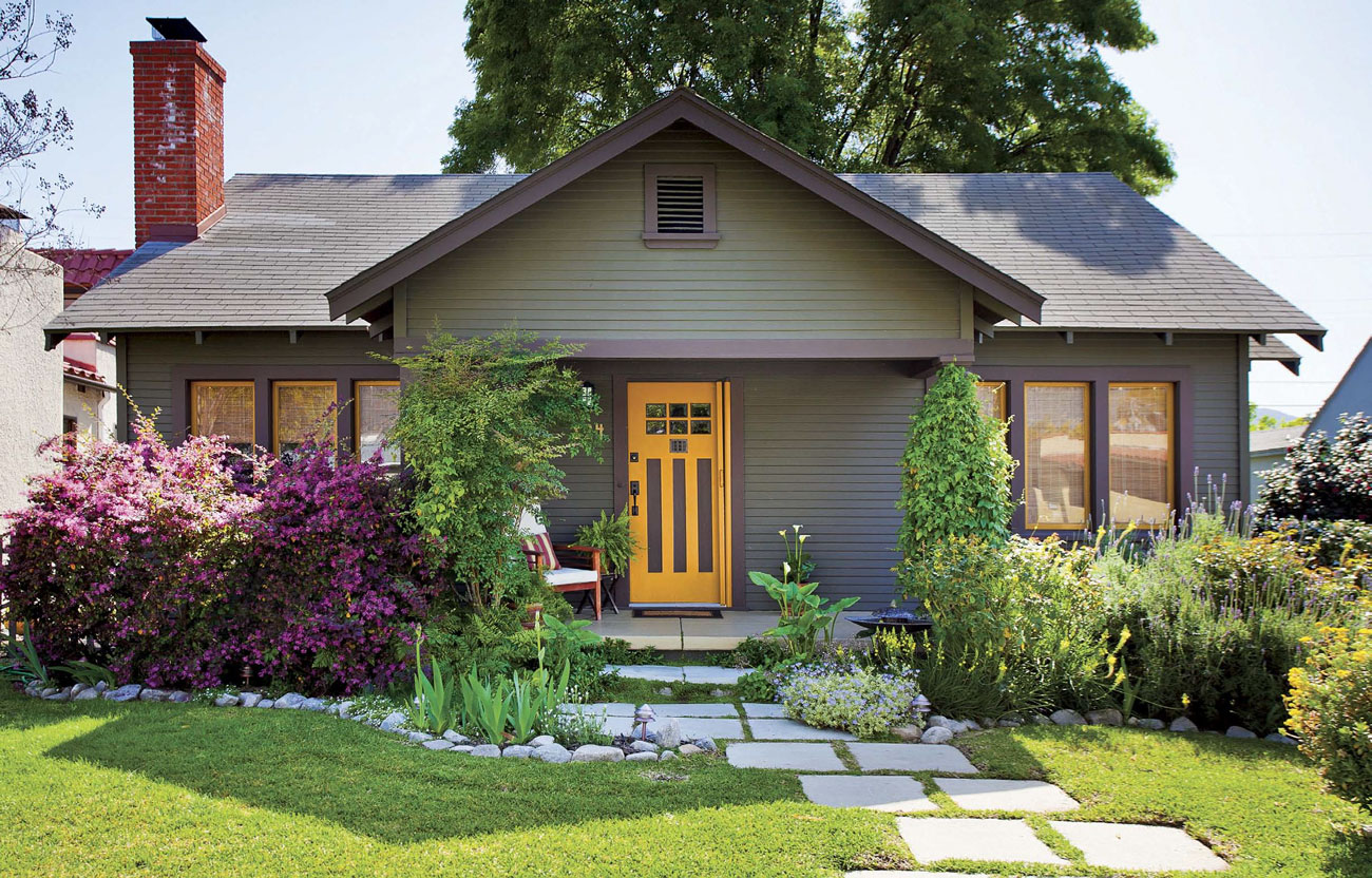 Home exterior of a muted green bungalow with stone pavers up to a bright yellow front door. 