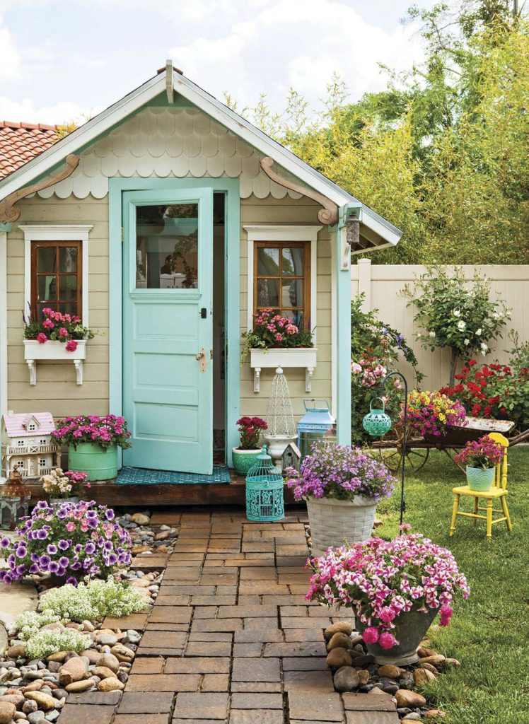 Vintage style playhouse in a yard surrounded by colorful potted flowers and a brick pathway.