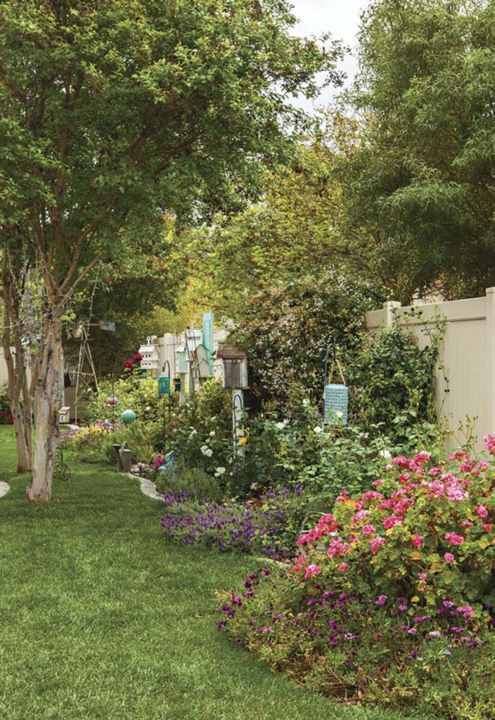 Planter filled with a variety of flowers, lanterns and birdhouses, running alongside a beige fence.