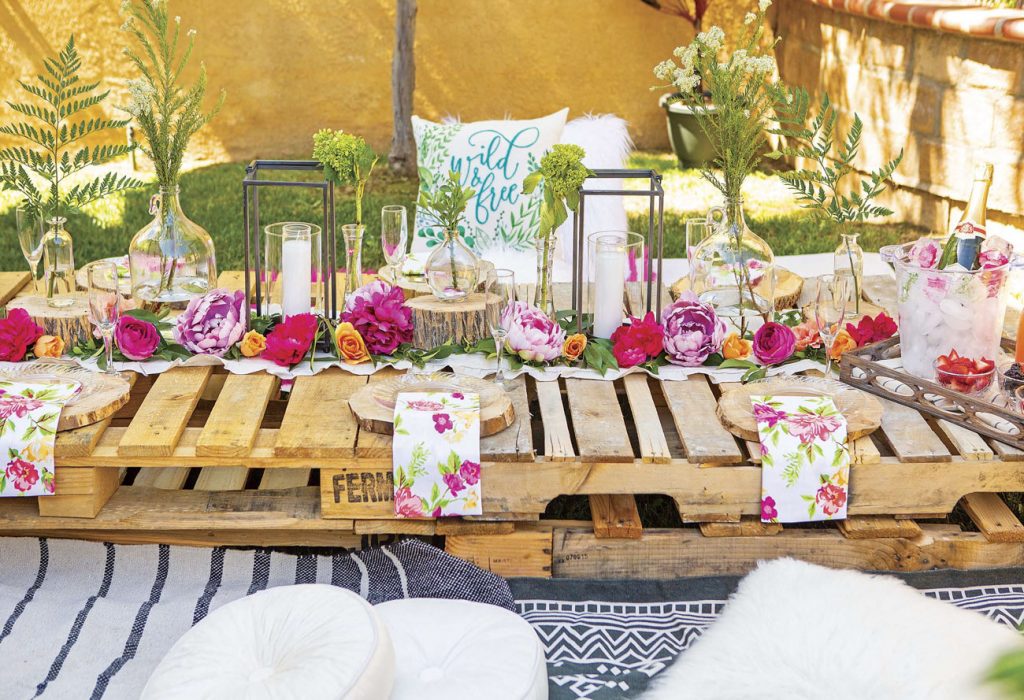 Garden party set up with a custom outdoor table made of wooden pallets and topped with bright pink and purple florals, greenery and lanterns. 