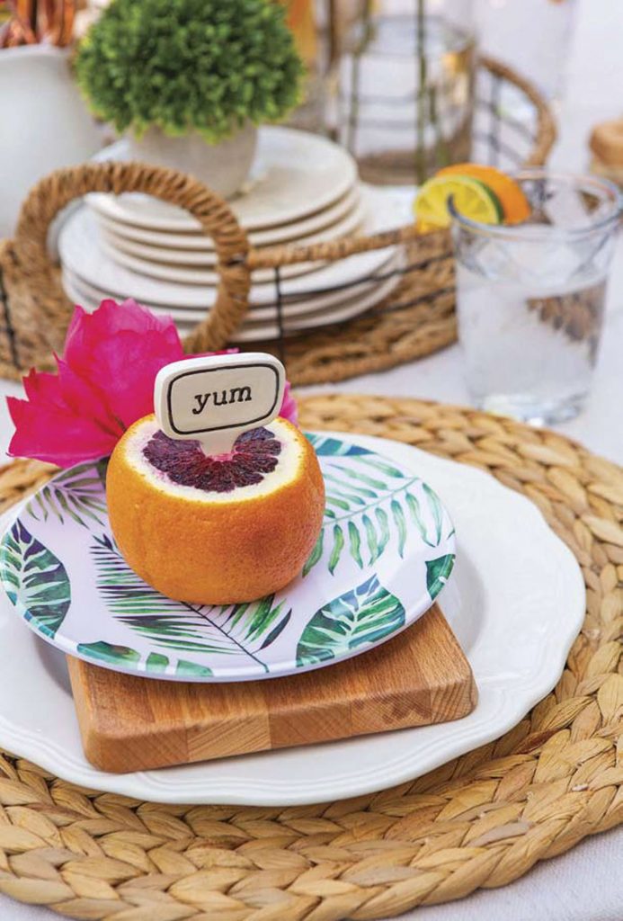 Garden party tabletop setting for a guest with woven charger plates and a blood orange and fresh fuchsia flower. 