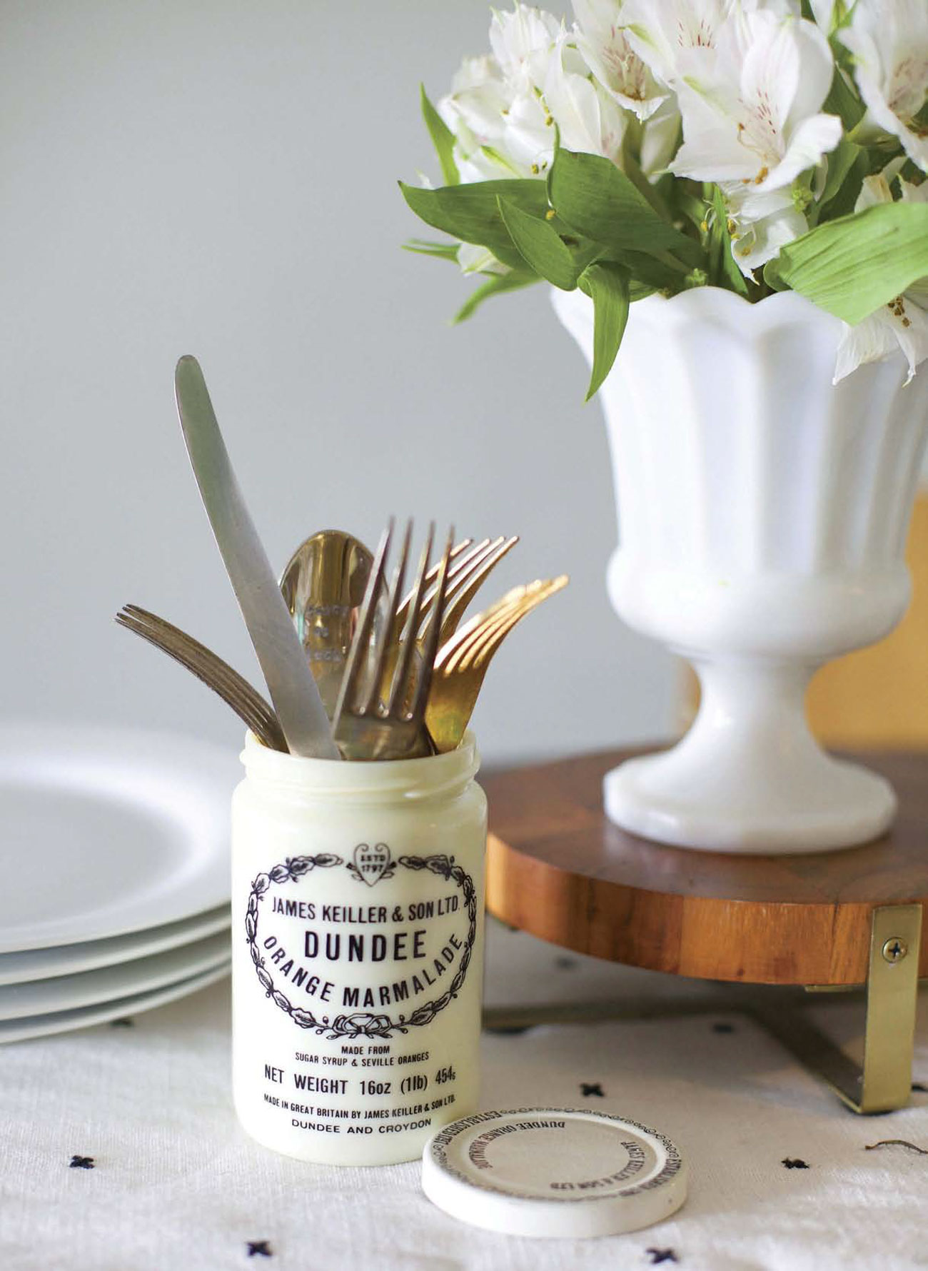 Keiller Dundee marmalade jar filled with silverware on a dining table next to white plates and a white vase of flowers.