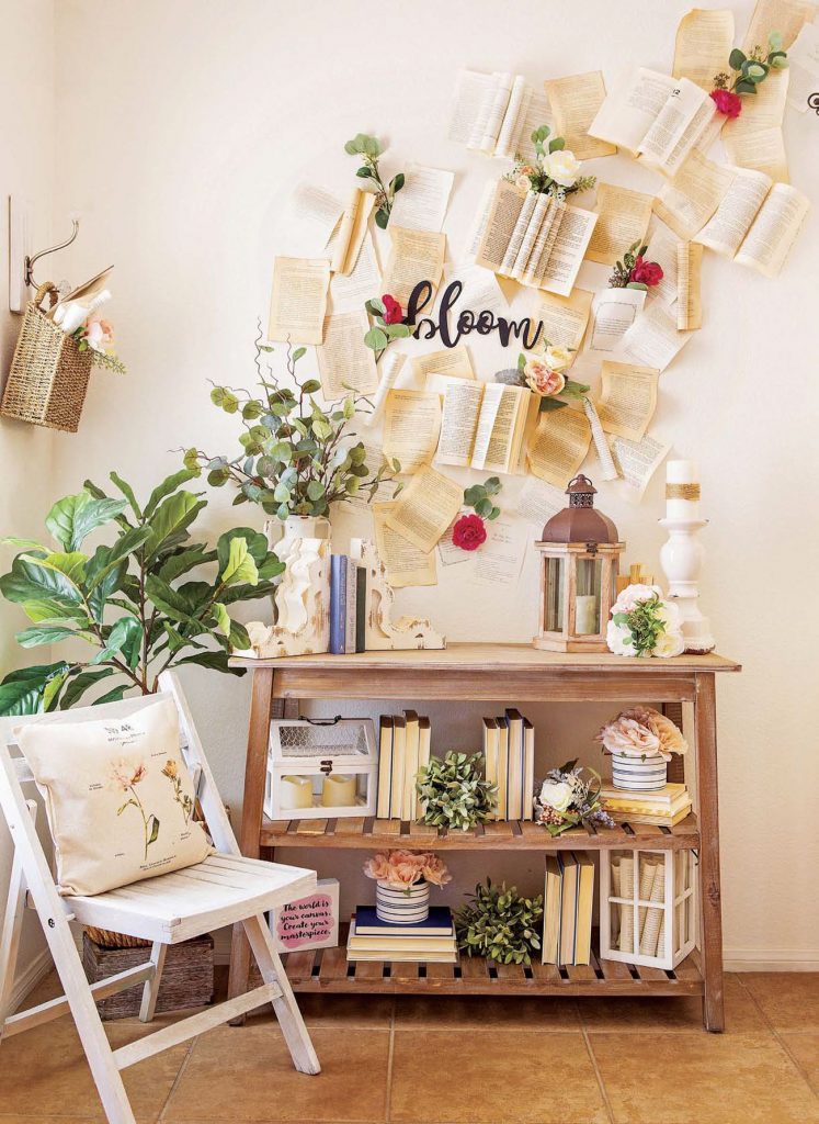 A folding, white wooden chair next to a magical looking "book wall" with seemingly flying books and pages making an art installation moving up the wall. 