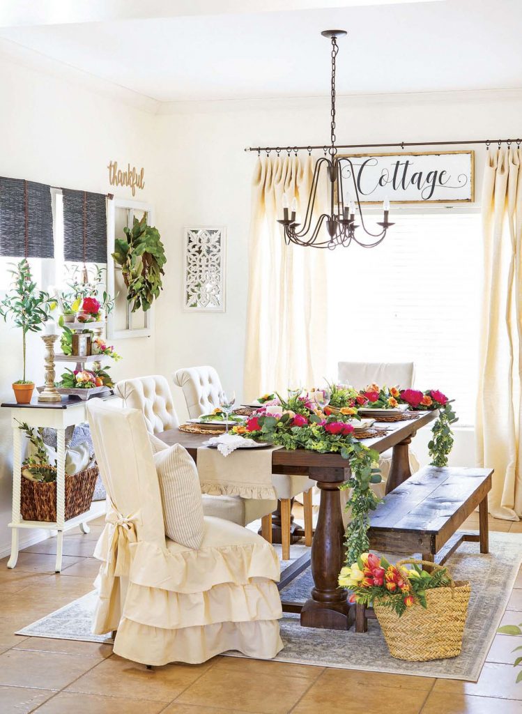 Cottage dining room with farmhouse table with wooden bench and fabric covered chairs surrounded by fresh flowers. 