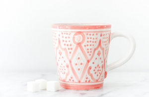 Ceramic white mug with pink designs wrapped around it sitting on a marble countertop next to sugar cubes.