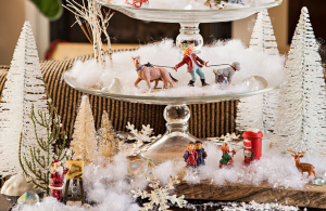 Glass cake stands stacked up with faux snow and small vintage Christmas figurines.