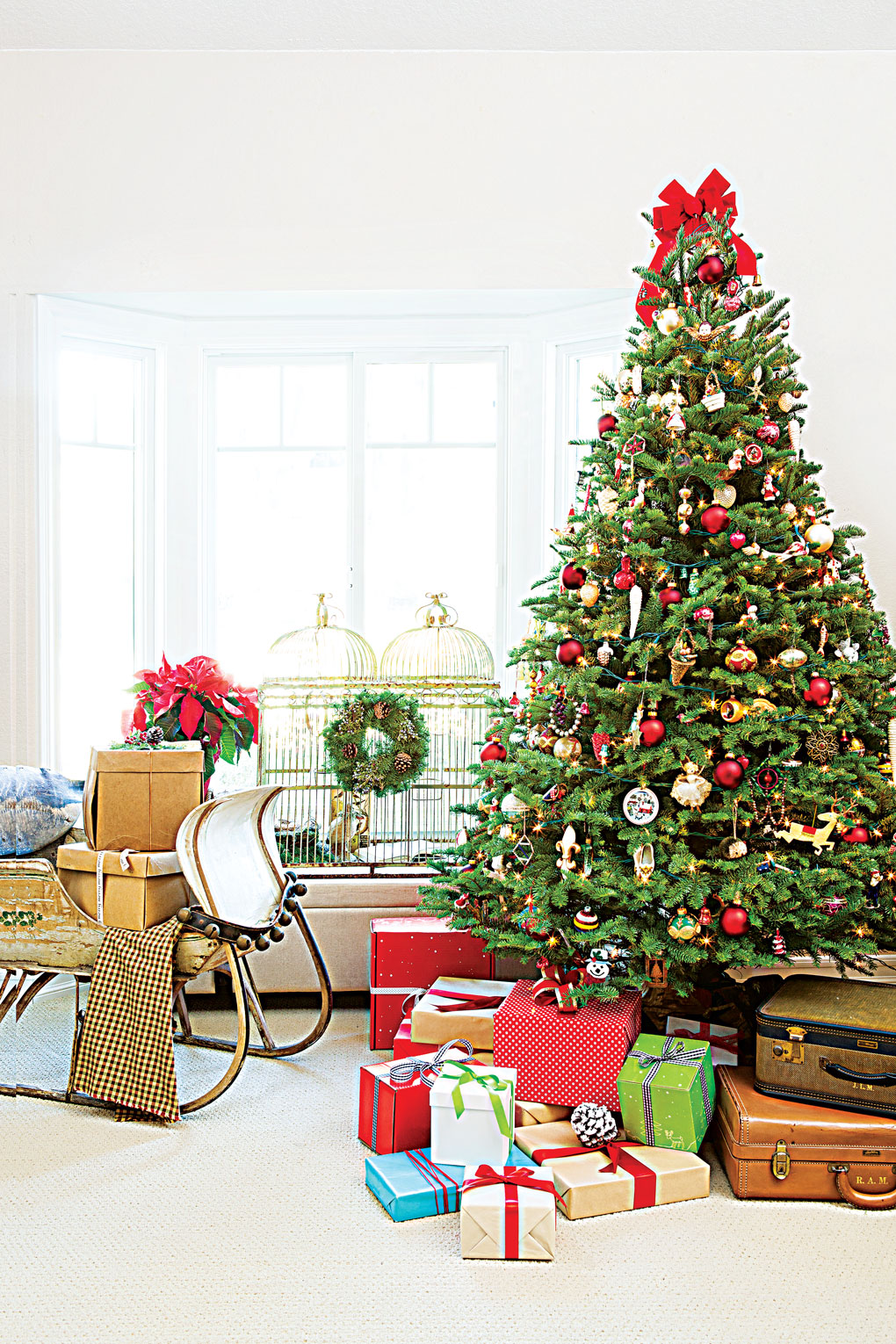 A large and lovely decorated Christmas tree topped with a red bow and surrounded by vintage suitcases and wrapped gifts all next to a vintage sleigh. 