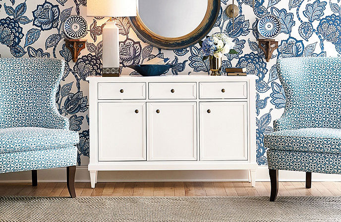 White sideboard placed in front of a blue floral wall with a large mirror above it and a white lamp on it.