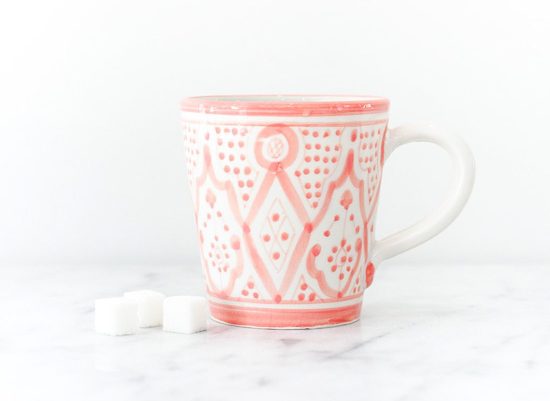 Ceramic white mug with pink designs wrapped around it sitting on a marble countertop next to sugar cubes. 