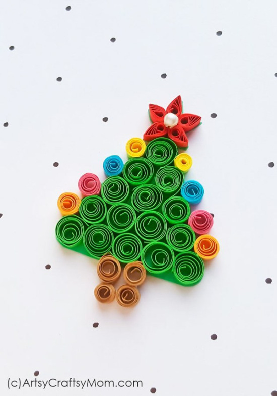 A bright colored paper quilled Christmas tree ornament laying flat on a white piece of paper with polka dots. 