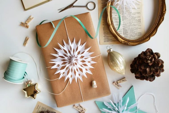 Smalle present wrapped in Kraft paper topped with white string and an ornate paper snowflake surrounded by teal accents, vintage frame, pinecone and golden ornaments. 