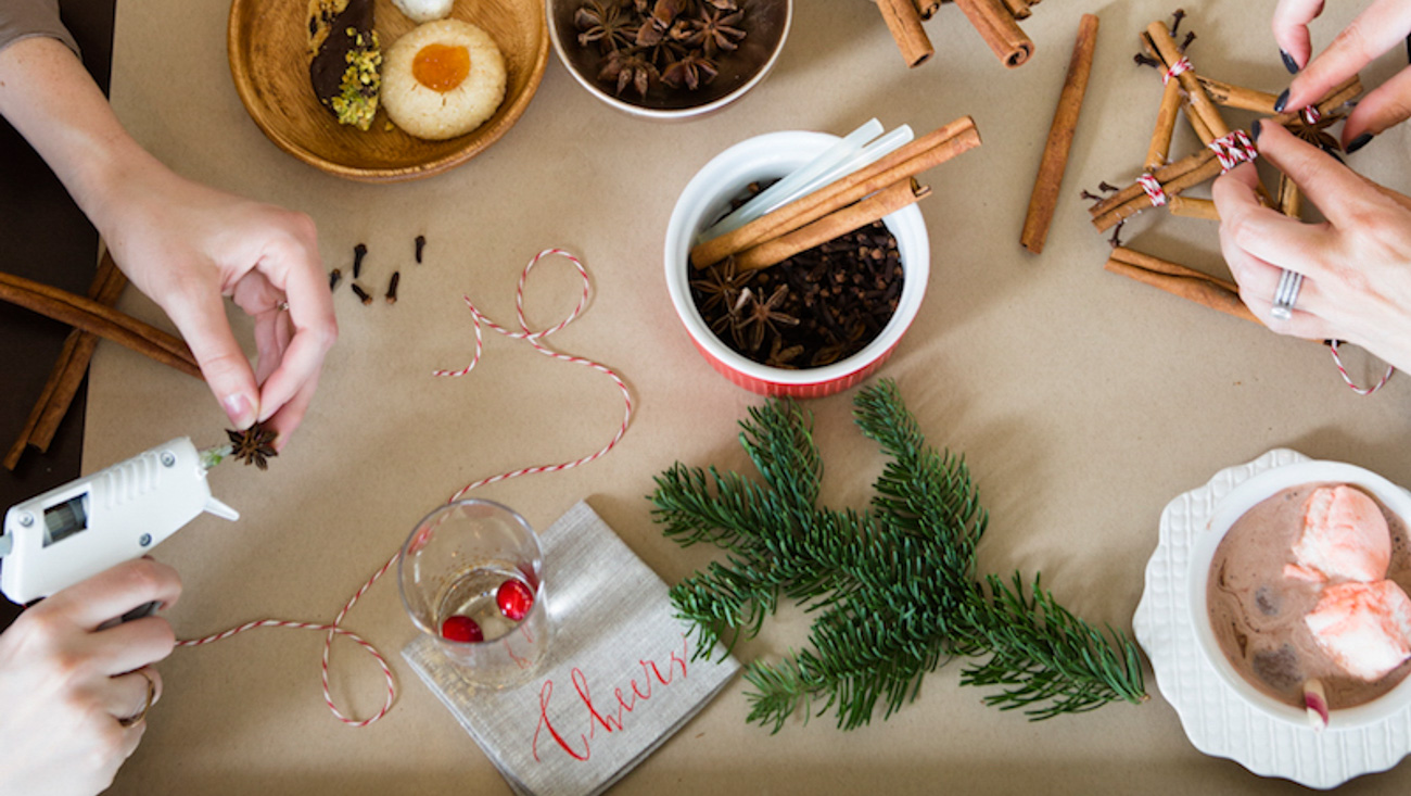 Overhead shot of hands crafting around a table with cups of cocoa, hot glue guns, shrubs and string.