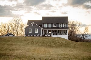 a dark greay farmhouse on a hill with woods behind