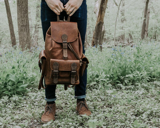 Man standing in the wilderness wearing dark denim and holding a leather backpack. 