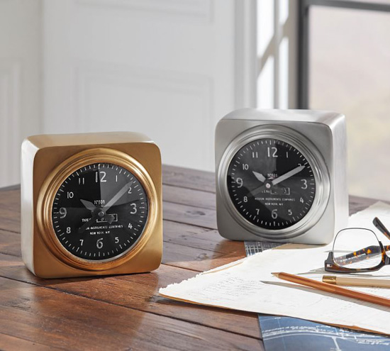 Two aviator alarm clocks now in chrome and one in gold placed on a wooden desk next to paper, pens and glasses. 