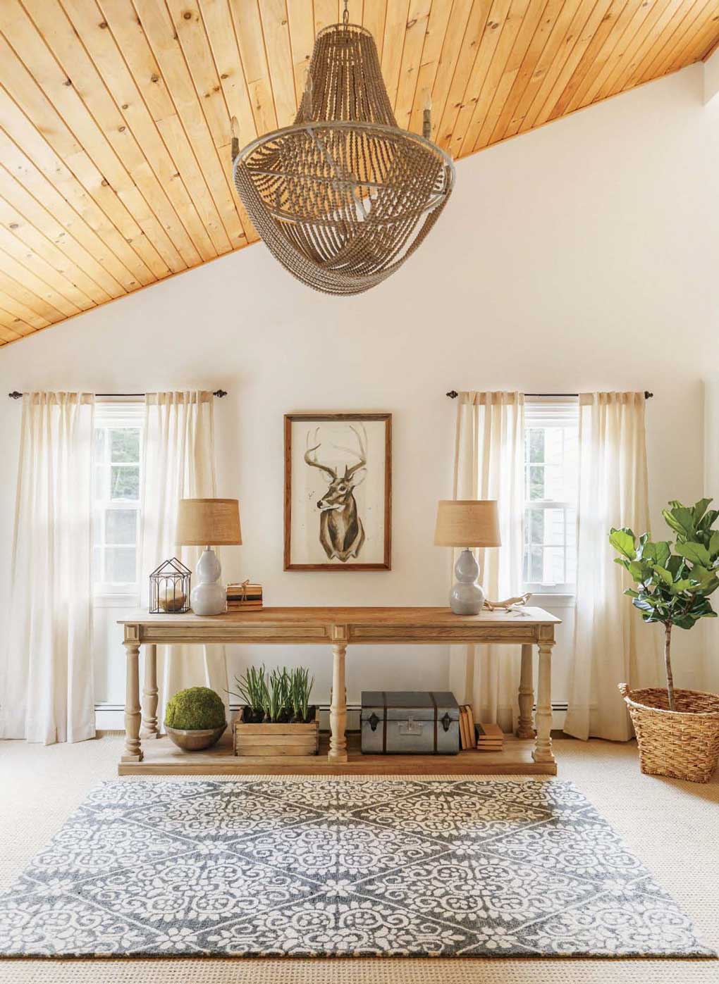 Rustic farmhouse open space with vaulted wooden ceilings and a beaded chandelier with a regal framed deer in between two windows. 