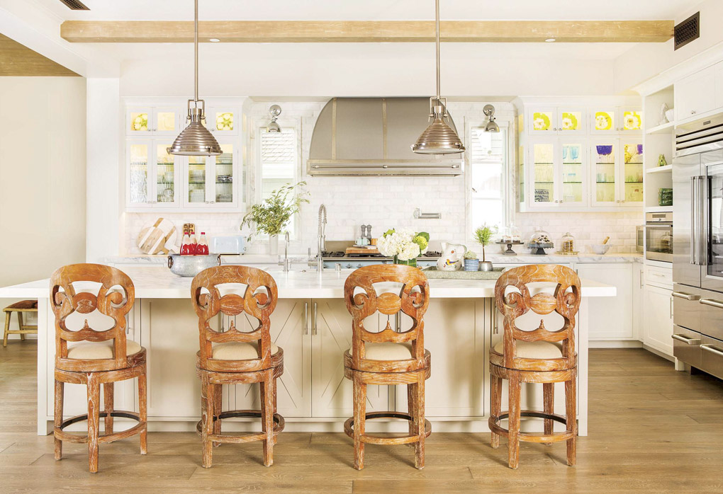 Wide open light colored kitchen looking over the backs of four uniquely carved wooden barstools.