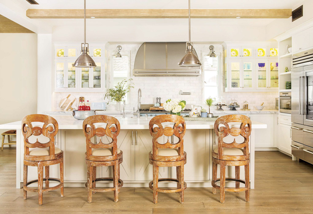 Wide open light colored kitchen looking over the backs of four uniquely carved wooden barstools. 