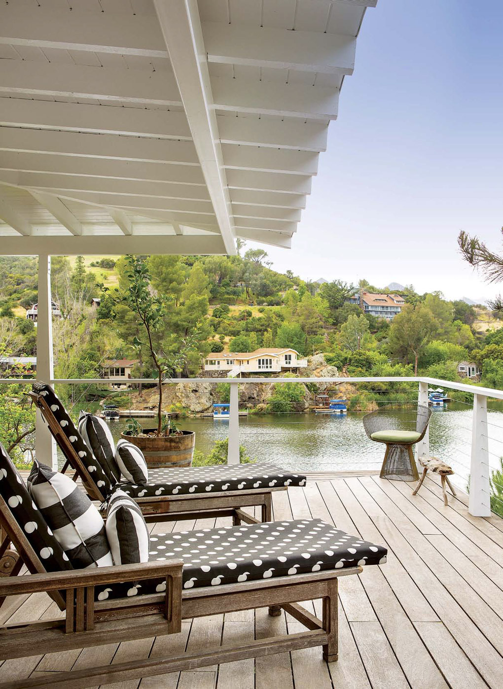 Teak lounge chairs with black and white cushions on a wooden outdoor deck overlooking a lake. 