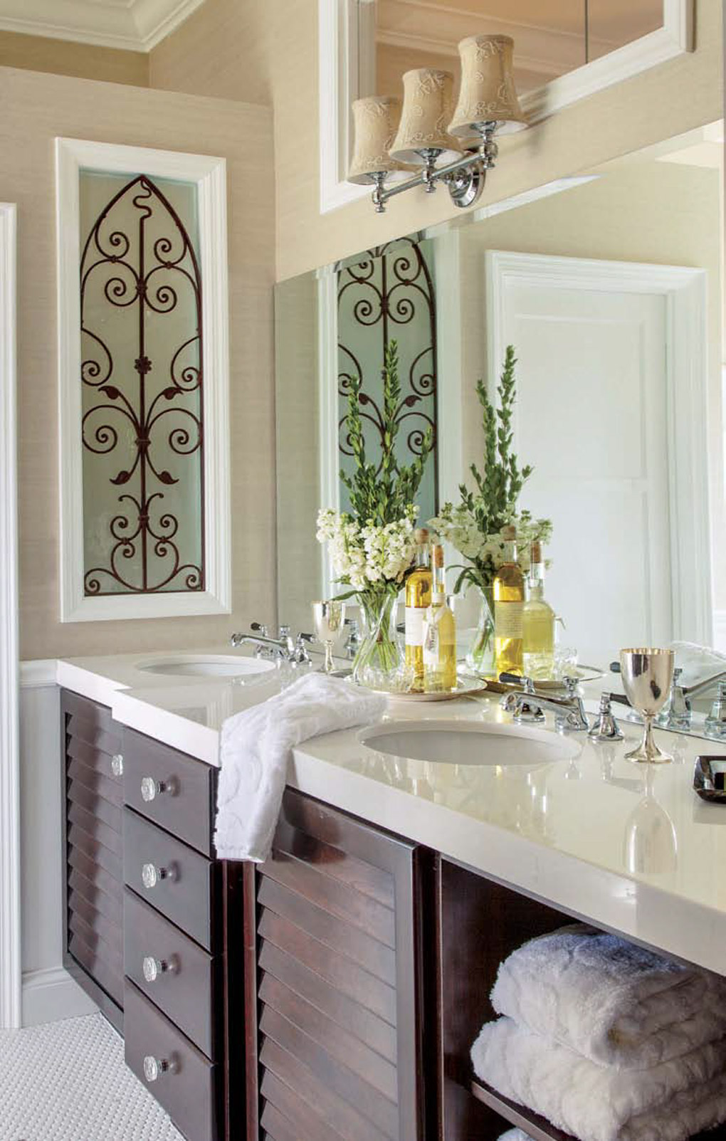 Double vanity bathroom sink area with dark wood drawers and tan accents on the wall and lighting with fresh flowers on the sink. 