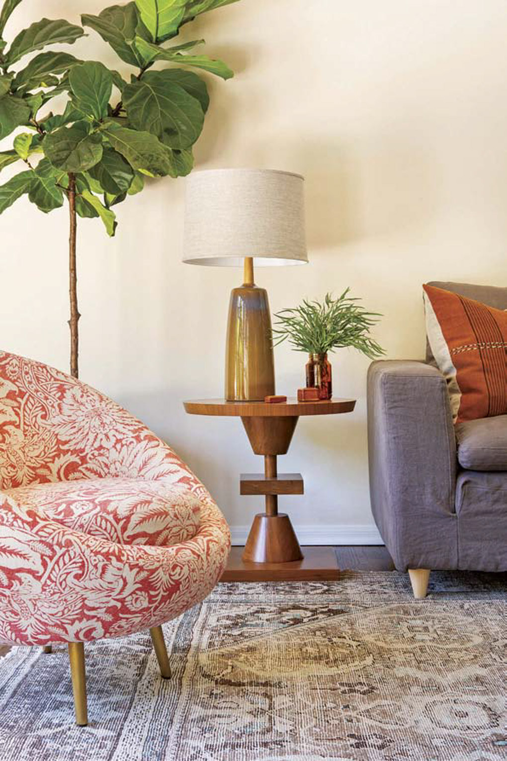 Modern side table made of walnut topped with a lamp and fresh greenery, next to a barrel-rolled floral chair and a gray couch. 