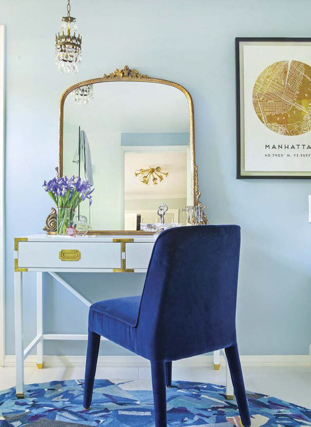 Bathroom vanity area with a gold accented mirror, hanging chandelier and a bright blue velvet chair. 