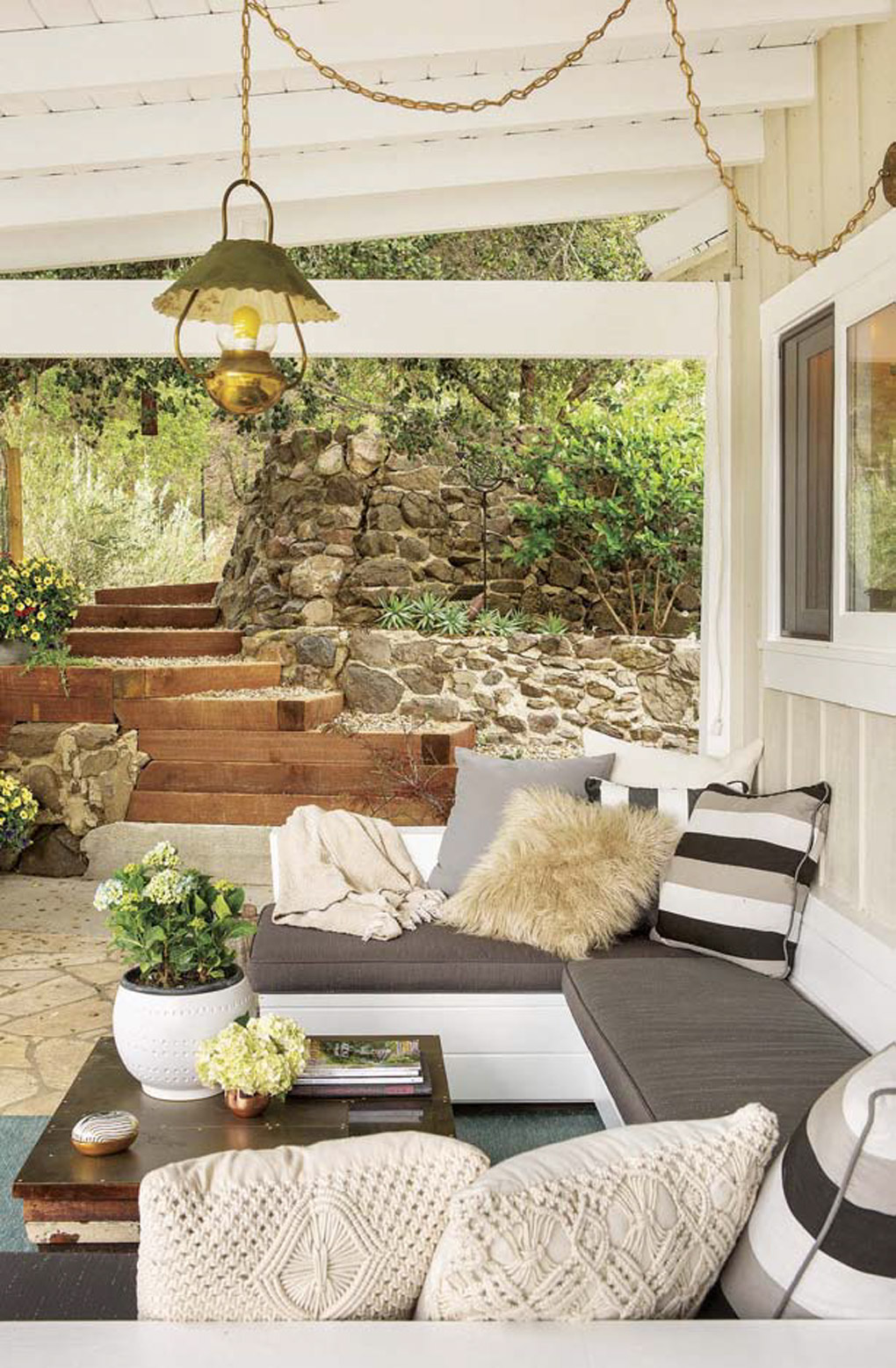 Hanging brass chained vintage light over an outdoor sectional with a vintage wooden coffee table and stone made walls with wooden steps in the background. 