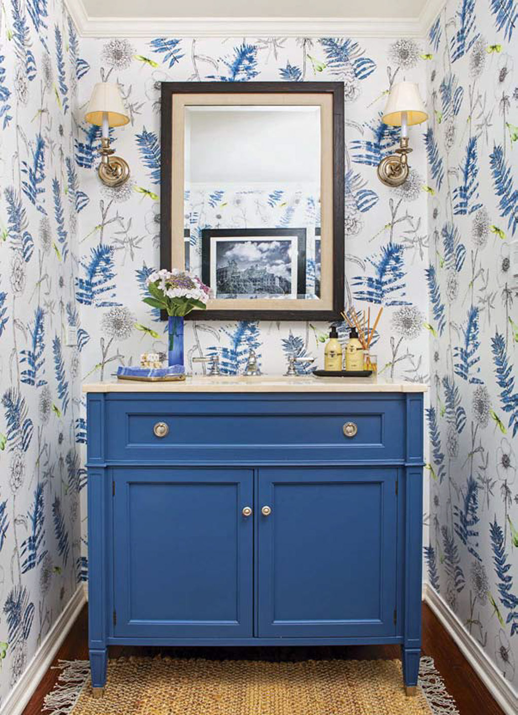 Powder bath with a bold blue vanity, surrounded by gold accents and floral wallpaper. 