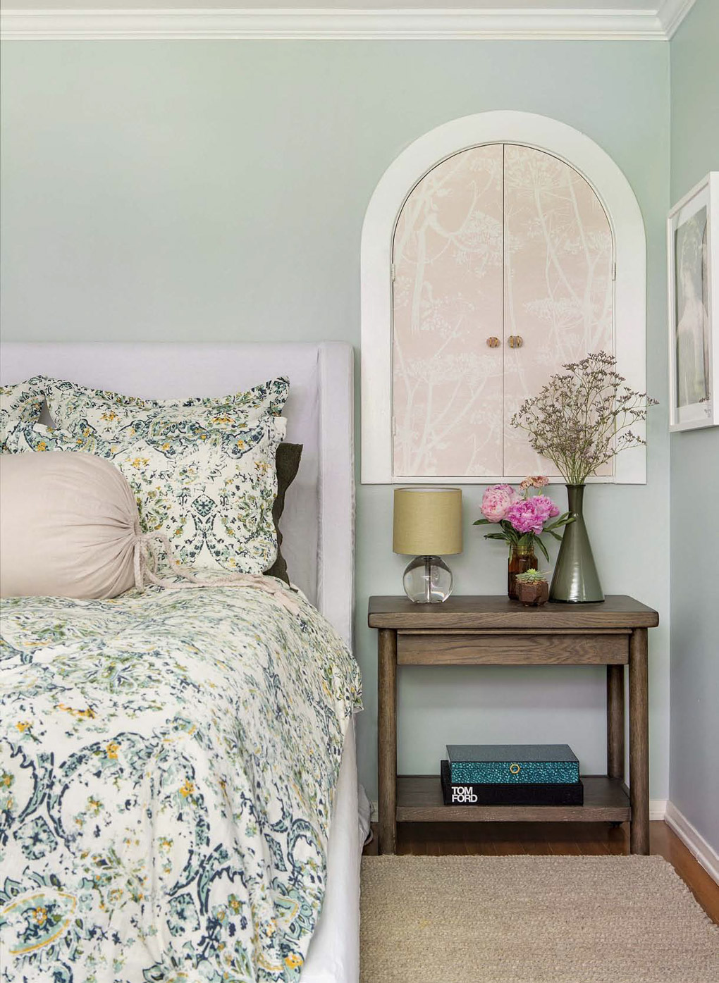 Floral bedspread with a wooden bedside table beneath a round topped storage cabinet in a California cottage master bedroom.