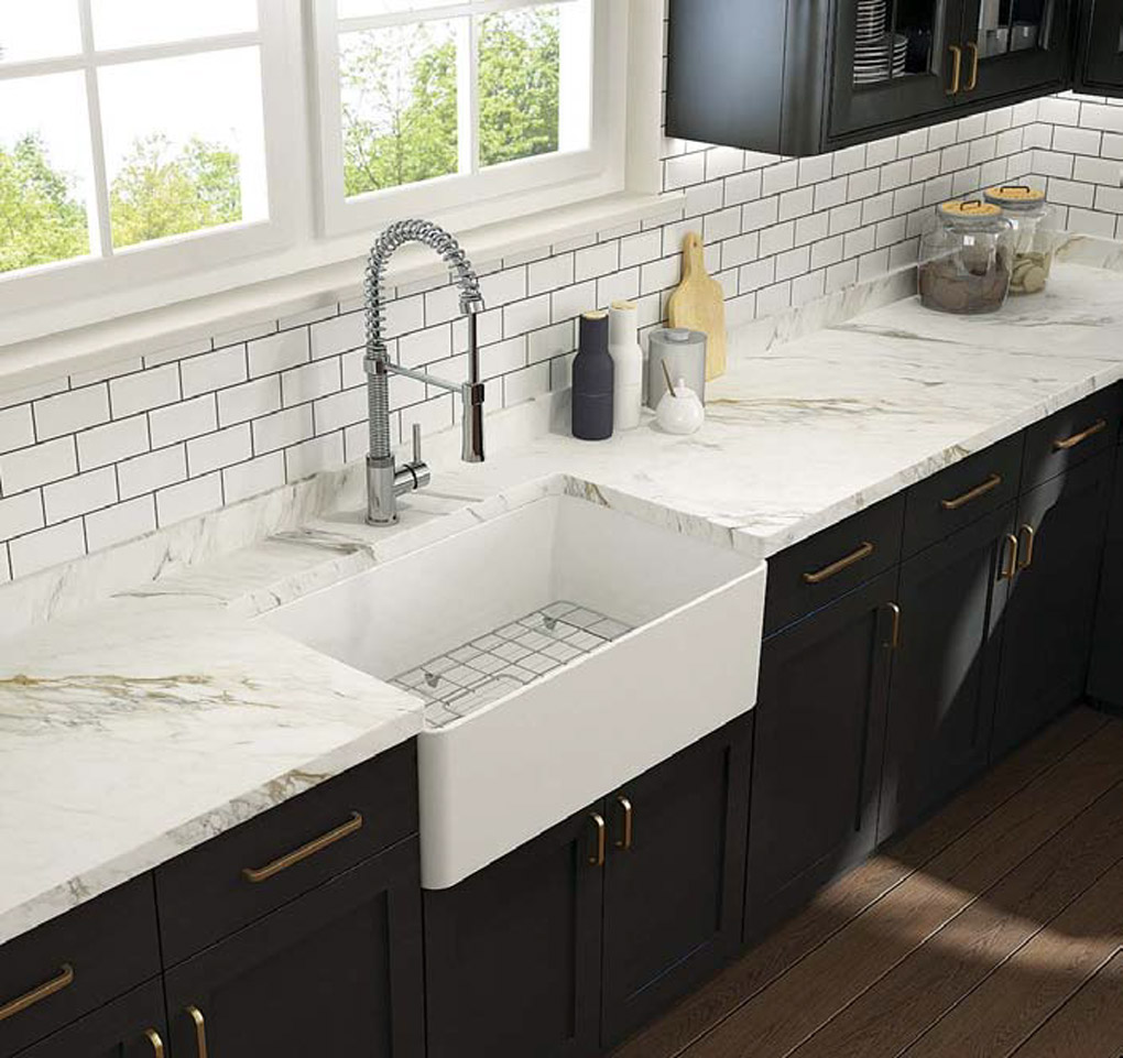 Farmhouse sink with marble counter tops in a kitchen with black cabinetry and brass handles. 