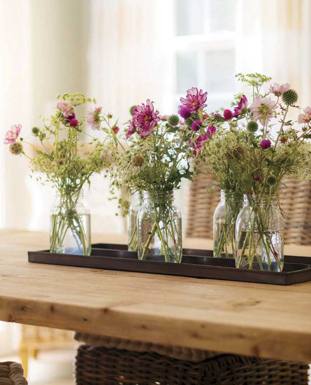 Mason jars filled with fresh-cut white and purple flowers placed together as a centerpiece on a farmhouse dining table. 