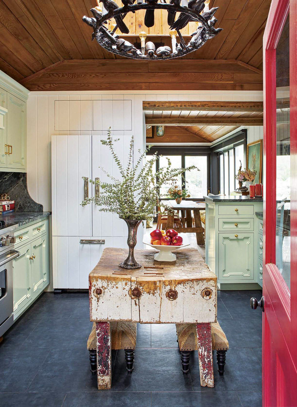 Bright red door opening into a cottage kitchen with rustic vintage island and light aqua cabinetry. 