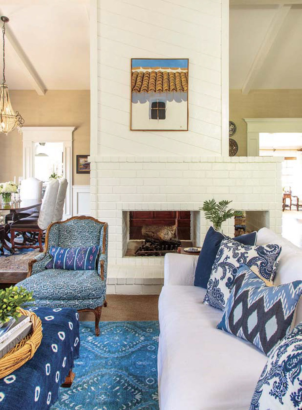 White, brick, walk-around fireplace, separating the dining from another seating area with blue rug, ottomoman and throw pillows accenting the white furniture. 