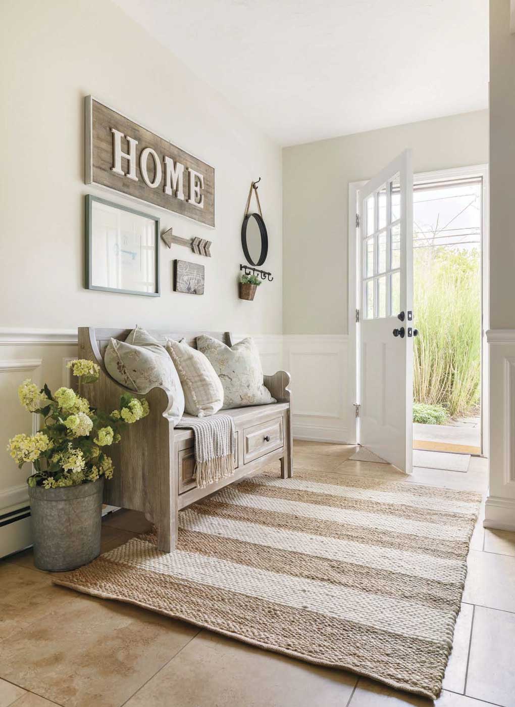 An open white door leading in to a casual mud room, with a light wooden bench, a wall sign that says "home". and a striped jute rug. 