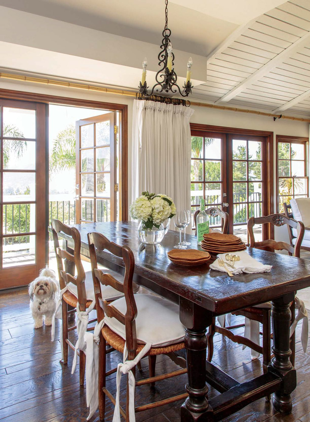 Antique French wooden truss table near a wall of double French doors with a small fluffy white pup looking on . 