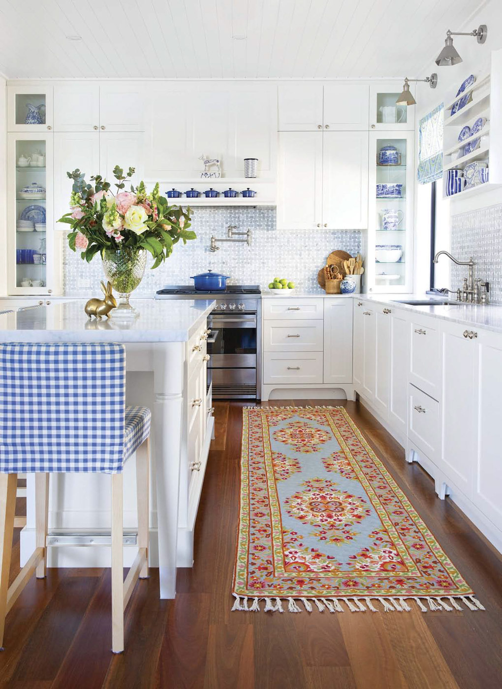 All white kitchen with cobalt blue accents and items in shelving, blue upholstered gingham bar stool and colorful floor runner. 