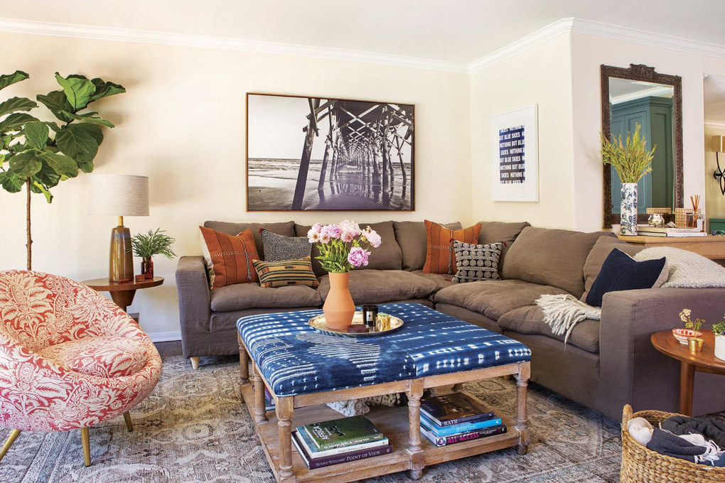 Wide shot of a living room in a California cottage with a floral barrel-back chair, sectional gray couch and a coffee table covered in Shibori fabric.