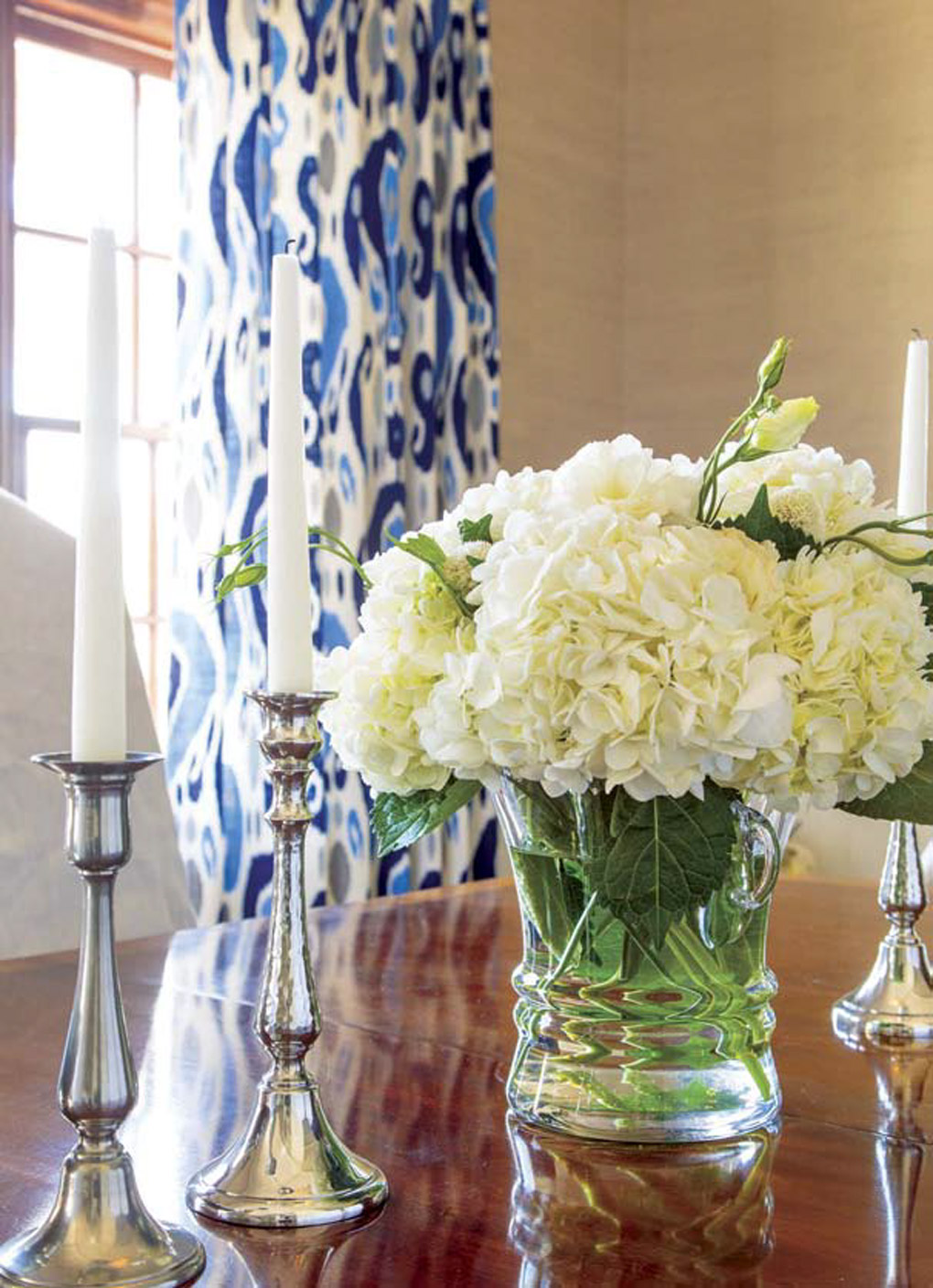 Full creamy hydrangea flowers in a clear vase on the table next to silver candlesticks on a wooden dining table with vibrant blue patterned curtains in the background. 