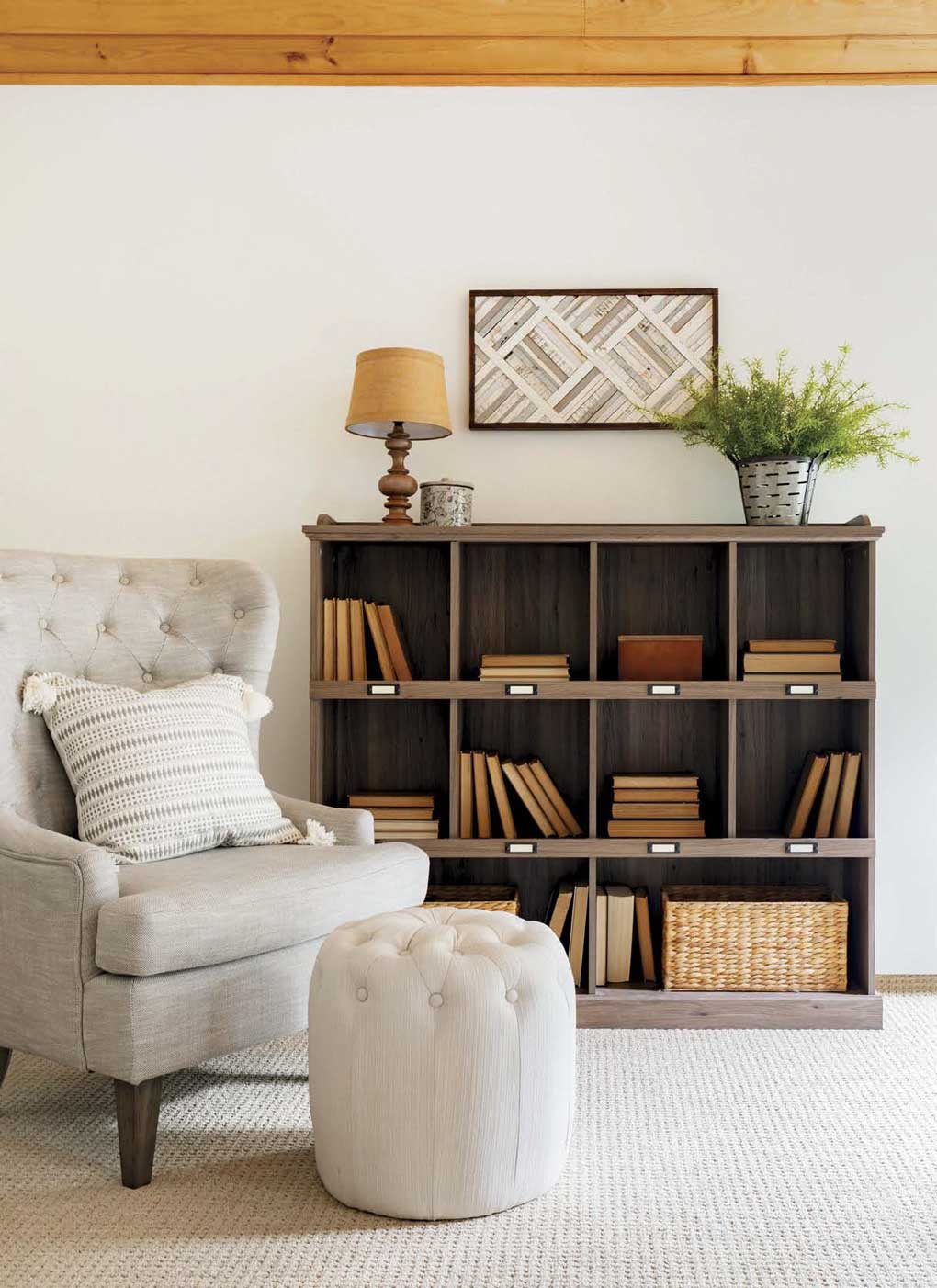 Heather gray tufted slip chair and matching foot rest next to a wooden bookshelf. 