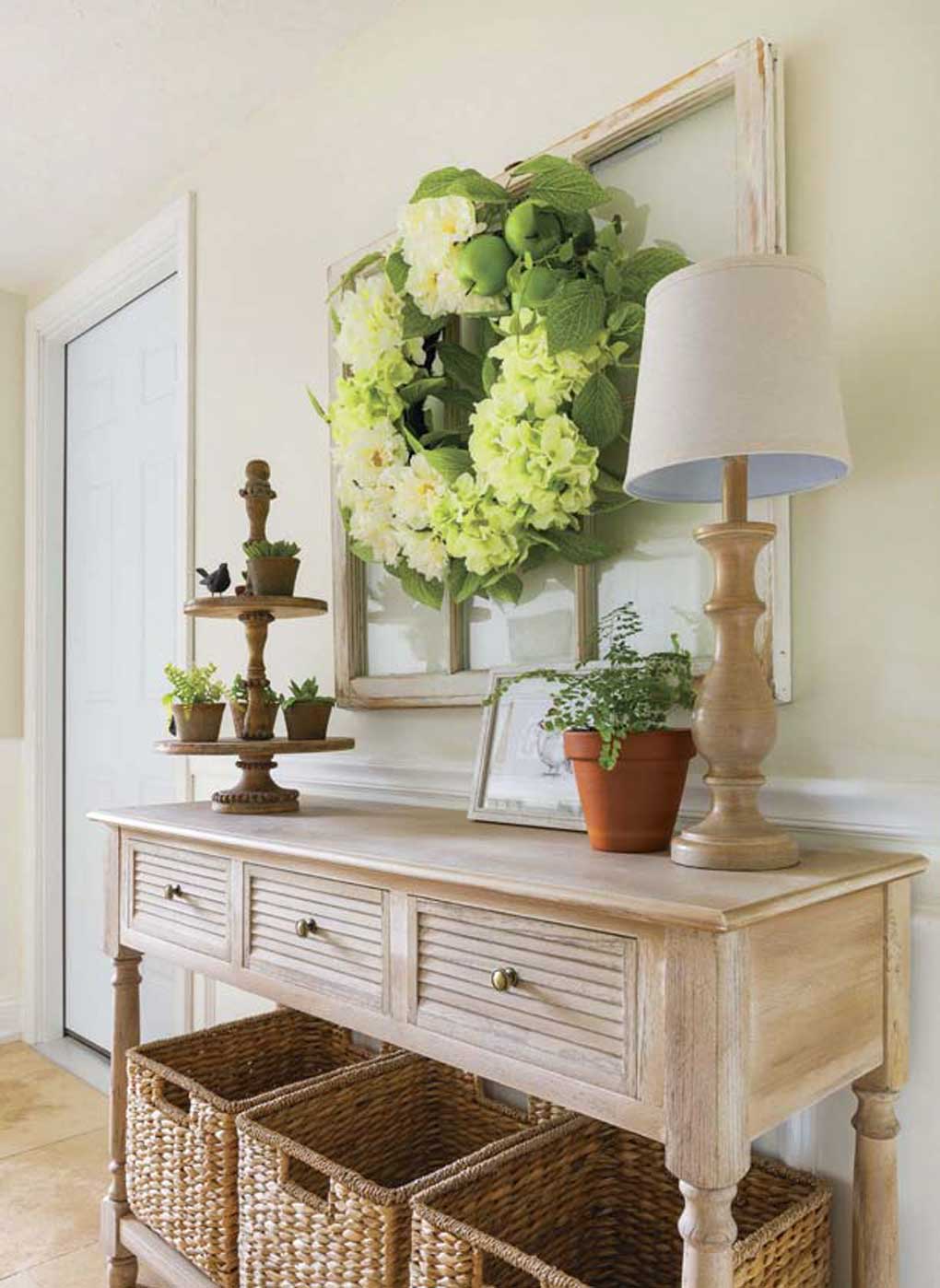 Entryway console table topped with plants and a lamp with three woven baskets underneath for storage as people enter the home and need to leave belongings. 