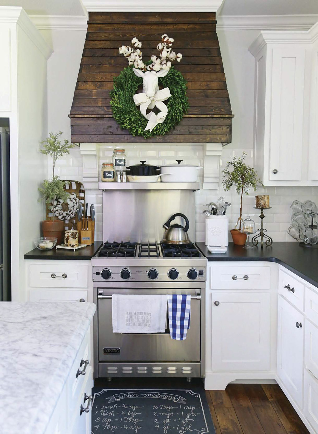 Kitchen with white cabinets and black countertops showing off a custom range hood with slatted wood and a bright green wreath with a white deer accent in the middle.