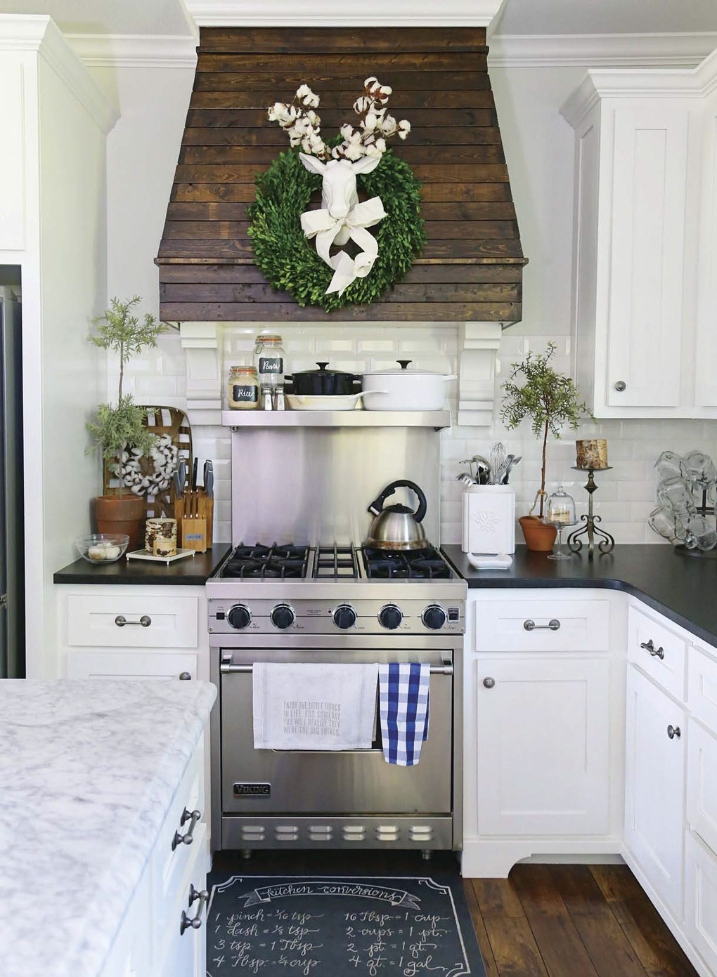 Kitchen with white cabinets and black countertops showing off a custom range hood with slatted wood and a bright green wreath with a white deer accent in the middle. 