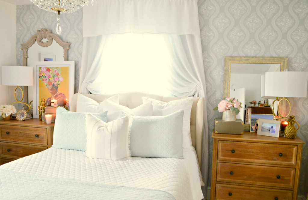 White and bright bedroom with pillows and window coverings flanked by two wooden dressers topped with art and lamps. 