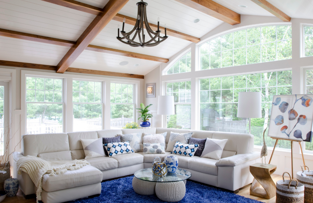 Modern cottage great room with exposed beams, classic blue rug and cream colored sectional surrounded by walls of windows. 